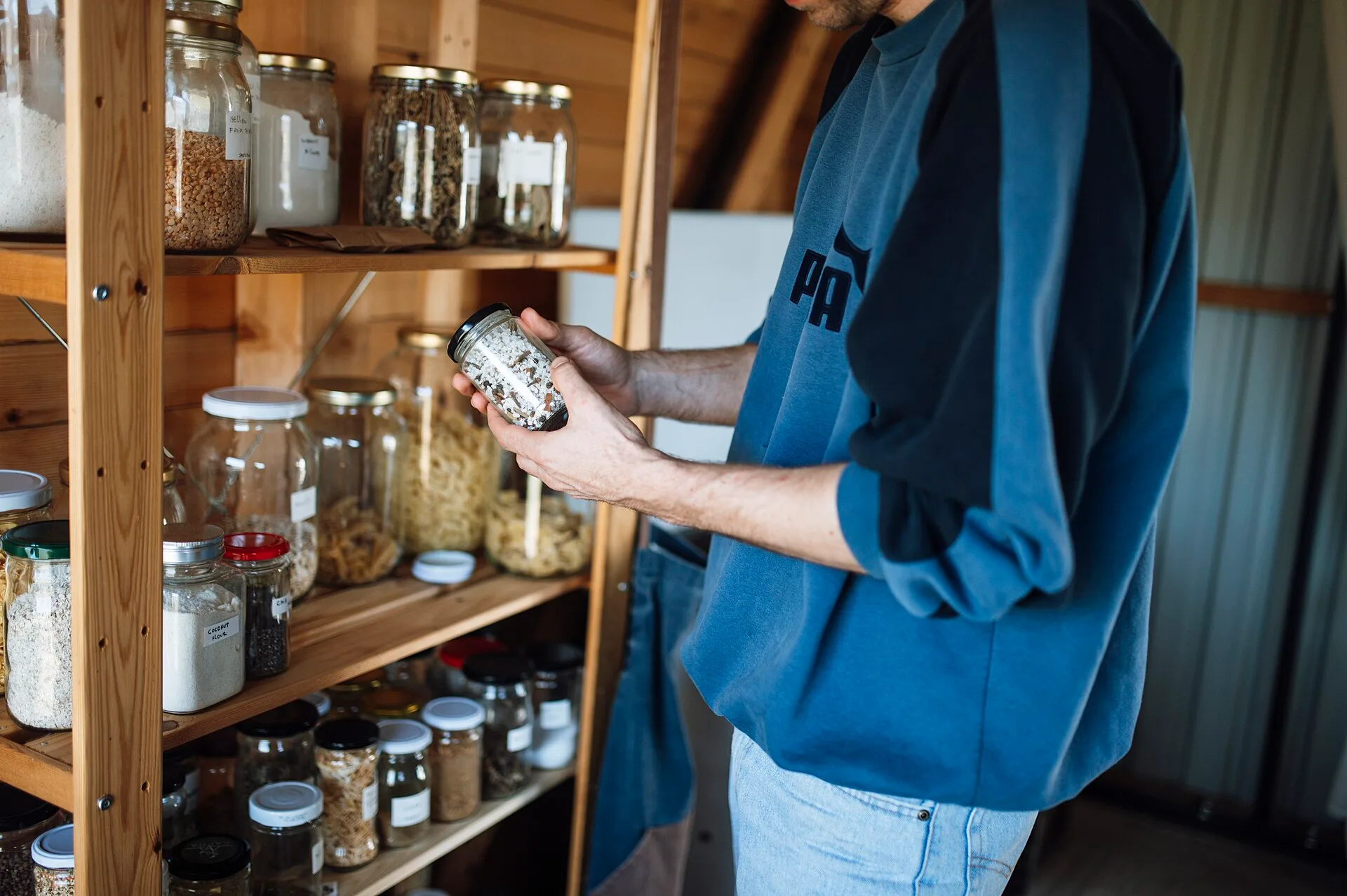 A person stands in a kitchen pantry looking closely at a jar filled with spices. The pantry has many jars arranged on wooden shelves. It is bright and organized. How to write up a year of pantry buying as a case study at home